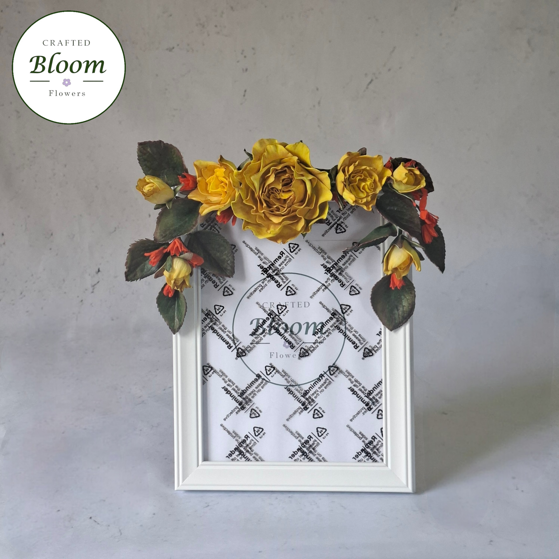 White picture frame decorated with yellow roses and green foliage on top. Crafted Bloom Flowers