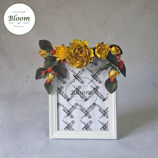 White picture frame decorated with yellow roses and green foliage on top. Crafted Bloom Flowers