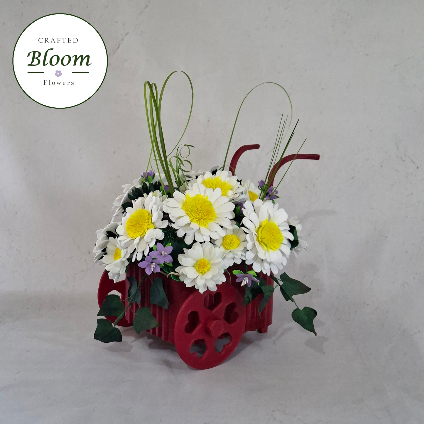 A floral arrangement in a red decorative wheel-shaped planter, featuring white daisies with yellow centers, green leaves, and small purple flowers, set against a light gray textured backdrop. A circular 'Crafted Bloom Flowers' logo is visible in the top left corner.