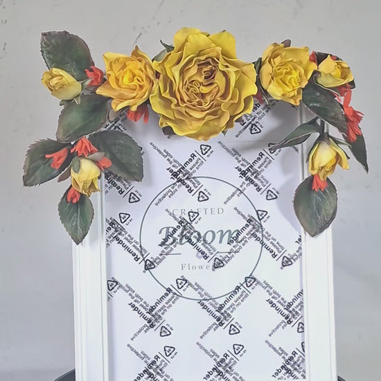 Detailed view of handmade yellow roses, rosebuds, and dark green leaves adoring a white frame corner